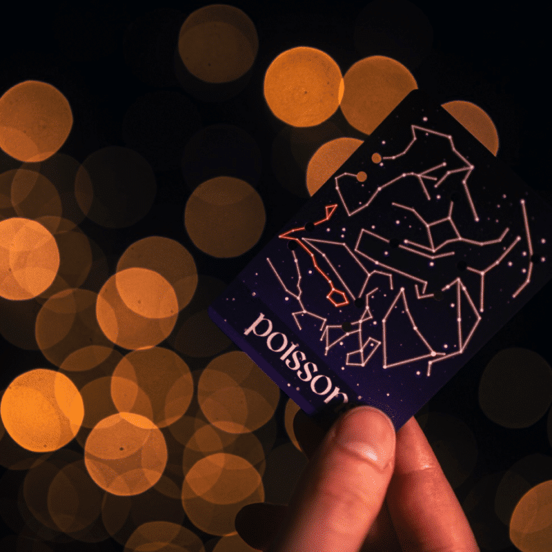 Une main affiche la carte « Constellations » sur un fond bokeh orange éclatant. La carte présente une illustration de constellation avec l'inscription « Poisson » en dessous, sur un fond sombre et rêveur.