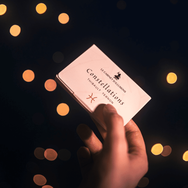 Une main tient un petit livre « Constellations » sur un fond sombre orné de lumières bokeh douces et lumineuses.