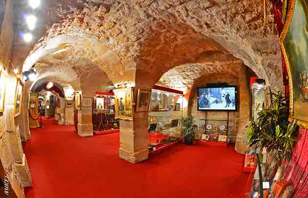 Une salle de musée confortable avec des arches en pierre et une moquette rouge. Des tableaux sont accrochés aux murs et des vitrines contiennent des objets. Un écran de télévision diffuse une image. Des plantes en pot ajoutent de la verdure à l'espace et l'éclairage est chaleureux et accueillant.